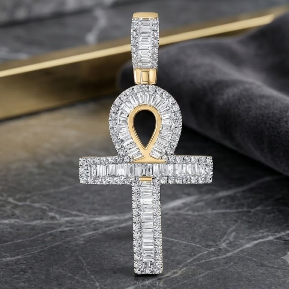 The 10K Gold Baguette Diamond Ankh Pendant 3/4 CTW rests on dark marble, its cross and loop shimmering with baguette-cut and round diamonds, while a gray fabric with a gold strip accents the background.