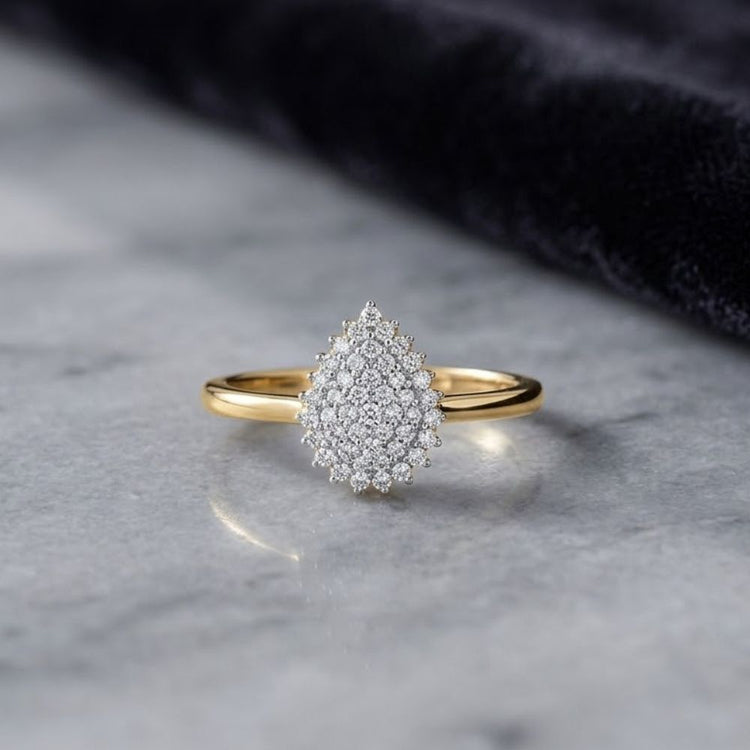 A beautiful 10k Yellow Gold 1/6CT Diamond Pear Gift Ring, featuring a teardrop-shaped cluster of sparkling diamonds, rests gracefully on a smooth marble surface with dark fabric in the background. This elegant ring makes the perfect thoughtful gift for someone special.