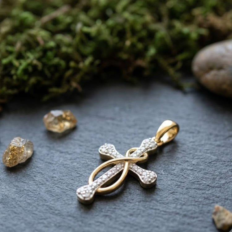 The 1/5CTW Diamond Cross Pendant in 10K Yellow Gold, featuring a looped design, is displayed on a dark stone surface with pieces of quartz and green moss around it.