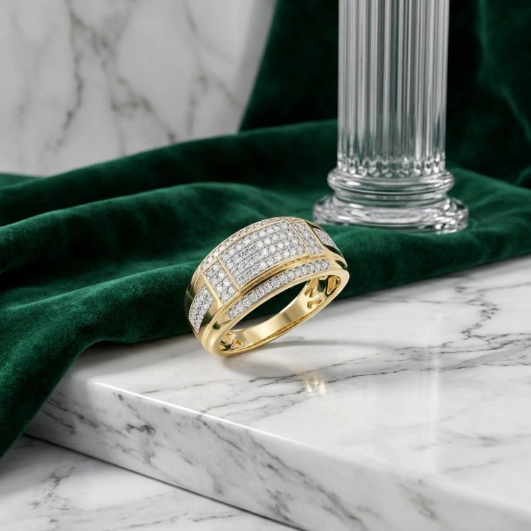 A 1/2CTW Natural Diamond Men’s Band in 10K yellow gold rests on a white marble surface, beside lush green velvet fabric and a glass column—a striking wedding band that radiates timeless elegance.