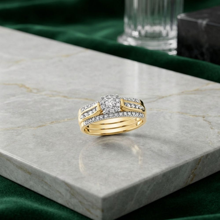 The 14k Yellow Gold 3-Piece Diamond Bridal Ring Set is beautifully displayed on a light marble surface, featuring multiple rows of sparkling diamonds. Green velvet fabric and a glass decoration in the background add an elegant touch to this exquisite presentation of diamond wedding rings.