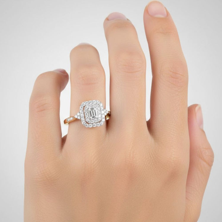 A hand with neatly manicured nails is wearing the 1/3CTW Diamond Emerald Double Halo Engagement Ring in 14k gold, featuring an emerald-cut diamond surrounded by two glittering halos of smaller diamonds.