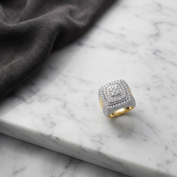 A striking 10K Yellow Gold Baguette Diamond Cluster Ring, adorned with sparkling diamonds in a square formation, rests on a marble surface beside folded dark fabric. This elegant piece effortlessly blends sophistication and style with its timeless yellow gold setting and radiant diamond cluster design.