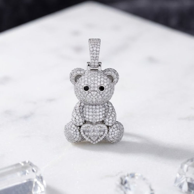 The Sterling Silver Moissanite Teddy Bear Heart Pendant features a beautifully crafted teddy bear with black eyes holding a heart, adorned with sparkling stones, and is displayed on a white marble surface beside blurred, shiny objects.