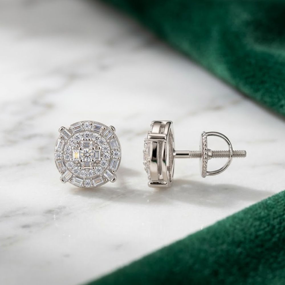 A pair of 925 Sterling Silver Baguette Round Moissanite Stud Earrings with screw-back posts is displayed on a white marble surface, partially bordered by green velvet fabric.