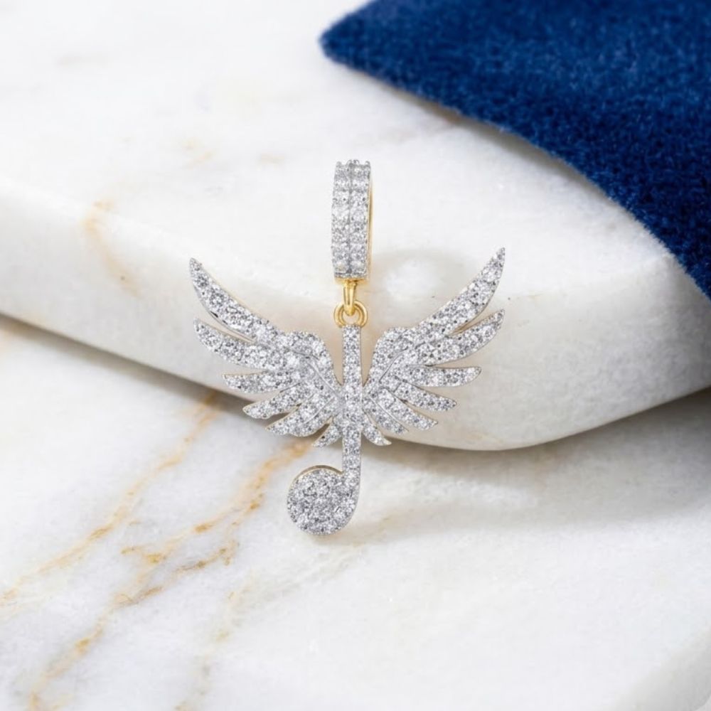 The 10K Gold Diamond Wing Music Note Pendant 1CTW, crafted in yellow gold with sparkling diamonds, is displayed on a white marble surface beside dark blue fabric.
