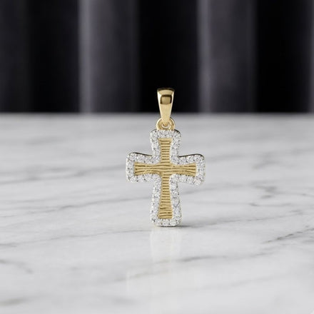 Showcased on a smooth white marble surface, the 1/5CTW Diamond Cross Pendant in 10K Yellow Gold features a textured center bordered by small, sparkling natural diamonds. The pendant’s radiant details stand out against a softly blurred background of dark, vertical drapes.