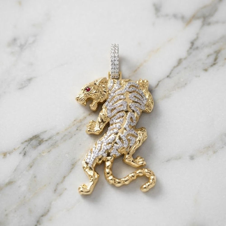 The Diamond Tiger Charm 1CTW 10K Yellow Gold features a diamond tiger with a red gemstone eye and a diamond-encrusted bail, displayed on white marble with grey veining.