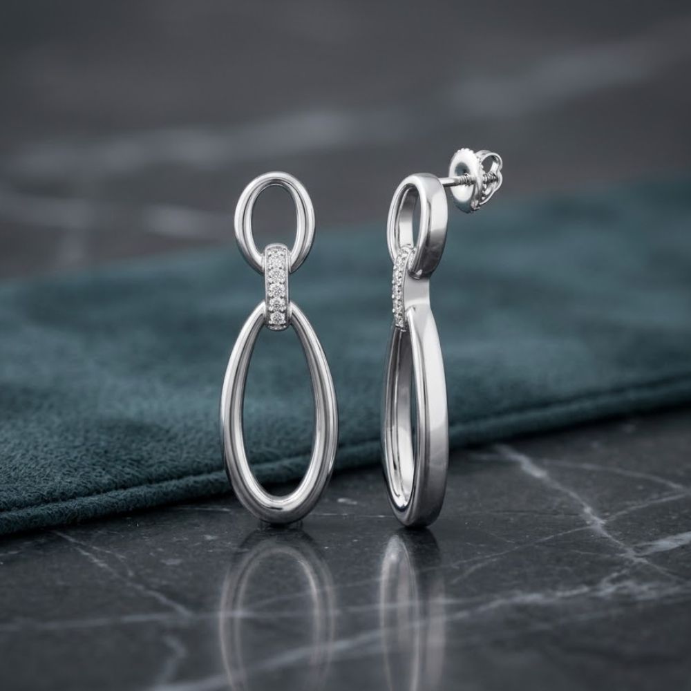 The 1/20 CTW Natural Diamond Dangling Earrings Silver feature two linked oval loops and sparkling natural diamonds, displayed on dark marble with a teal fabric backdrop. An elegant gift choice for any occasion.