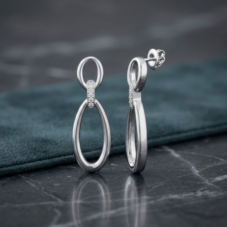 The 1/20 CTW Natural Diamond Dangling Earrings Silver feature two linked oval loops and sparkling natural diamonds, displayed on dark marble with a teal fabric backdrop. An elegant gift choice for any occasion.