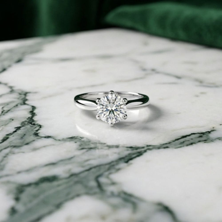 A 14k White Gold Round Diamond Solitaire Bridal Ring 1Cttw rests on a white marble surface with dark green veins, set against a blurred green background, highlighting the timeless elegance of this classic diamond engagement ring.