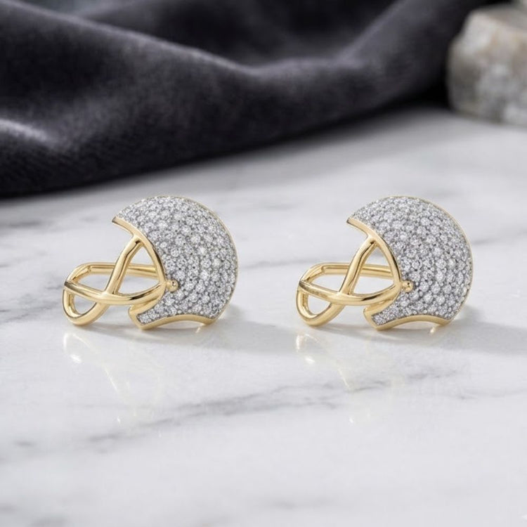 A pair of 3/8CTW Round Diamond Football Helmet Fashion Stud Earrings in 10K yellow gold, adorned with sparkling diamonds, are elegantly displayed on a white marble surface with a dark fabric backdrop.