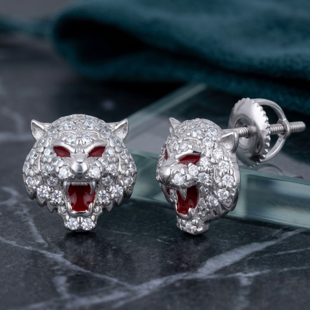 Close-up of Sterling Silver Moissanite Tiger Stud Earrings featuring roaring tiger heads with red gemstone eyes and open mouths, encrusted with sparkling stones, displayed on marble with a green fabric background.