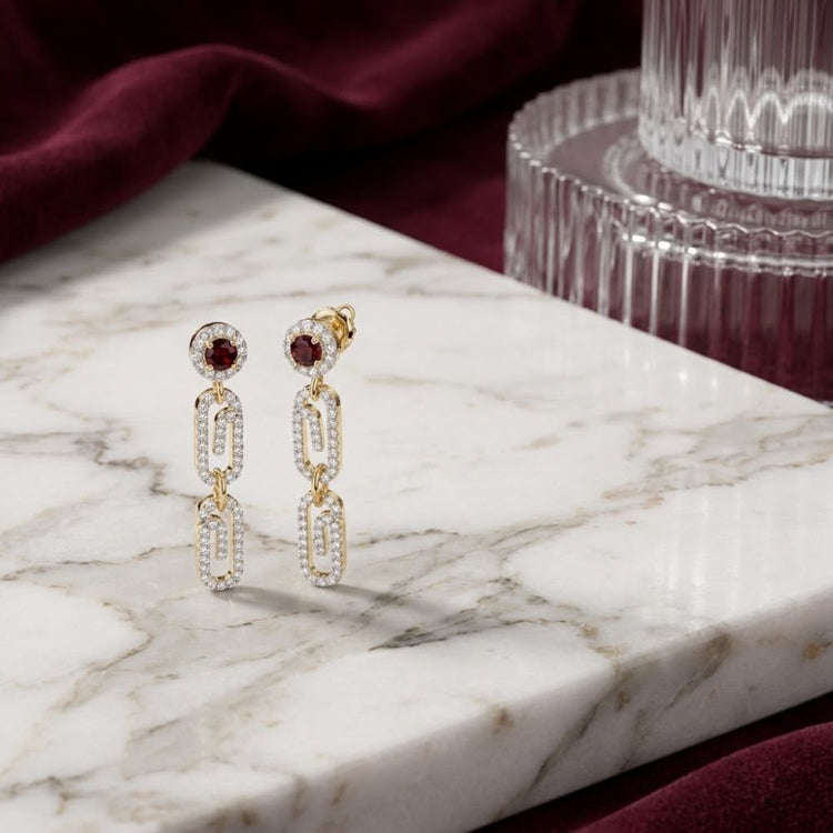 Displayed on a white marble slab, with burgundy fabric and a glass decor piece in the background, are the 1/2 CTW Natural Diamond Paper Clip Earrings. These elegant dangle earrings are crafted in 10K yellow gold and feature natural diamonds set in contemporary paper clip links for a look that is both modern and timeless.
