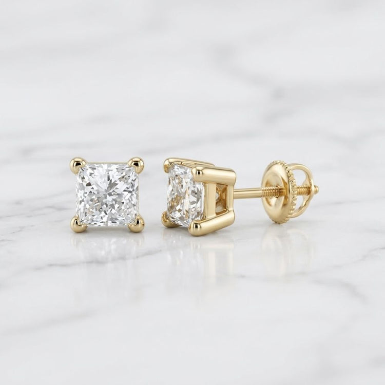 A pair of 14k Yellow Gold Princess Diamond Stud Earrings displayed on white marble; one stands upright and the other lies on its side, showing the screw-back closure.