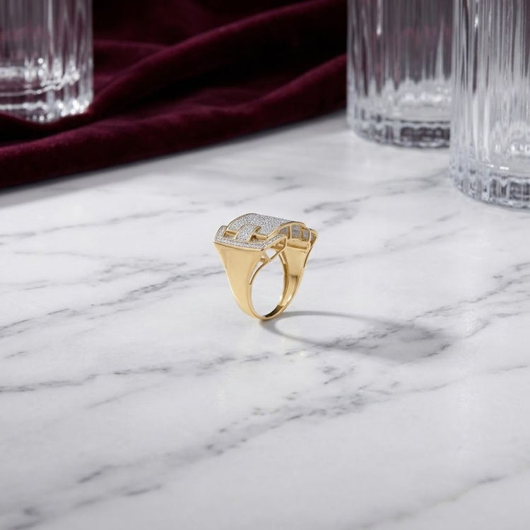 A captivating 10K Yellow Gold Diamond Cluster Ring 5/8 CTTW, featuring a diamond-encrusted square top, rests elegantly on a white marble surface, surrounded by clear glass tumblers and a burgundy cloth in the background.