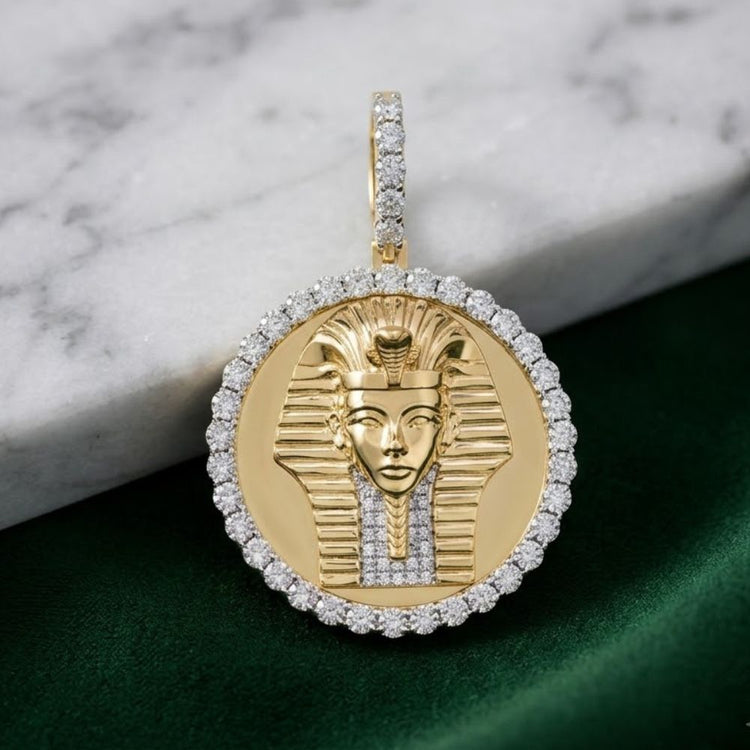 The Egyptian Face Diamond Pendant 1/4 CTW Silver is a round pendant crafted in yellow sterling silver, showcasing the visage of an ancient pharaoh surrounded by natural diamonds and displayed on a green cloth beside a white marble surface.