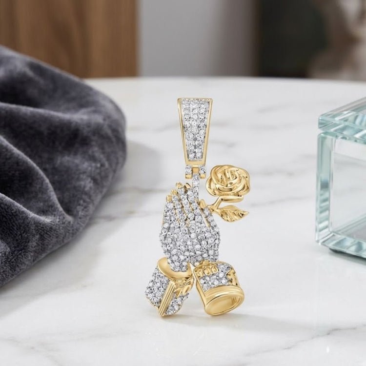 The Diamond Praying Hands Rose Charm 1/2CT 10K Gold features two praying hands holding a rose, displayed upright on white marble with a soft gray cloth and glass box, creating a striking praying hands necklace.