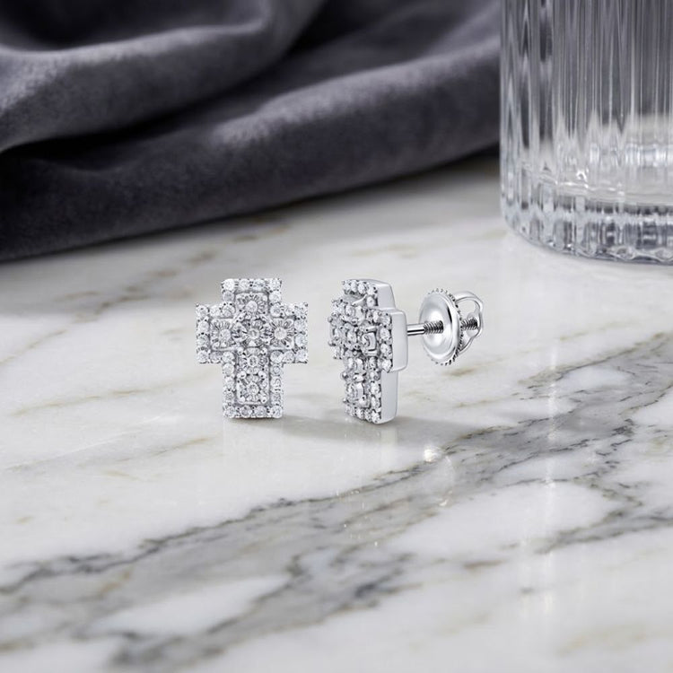 1/3 CTW Natural Diamond Cross Earrings Silver are showcased on a white marble surface, with shimmering natural diamonds accenting the sterling silver crosses. A textured gray cloth and a clear glass grace the background.