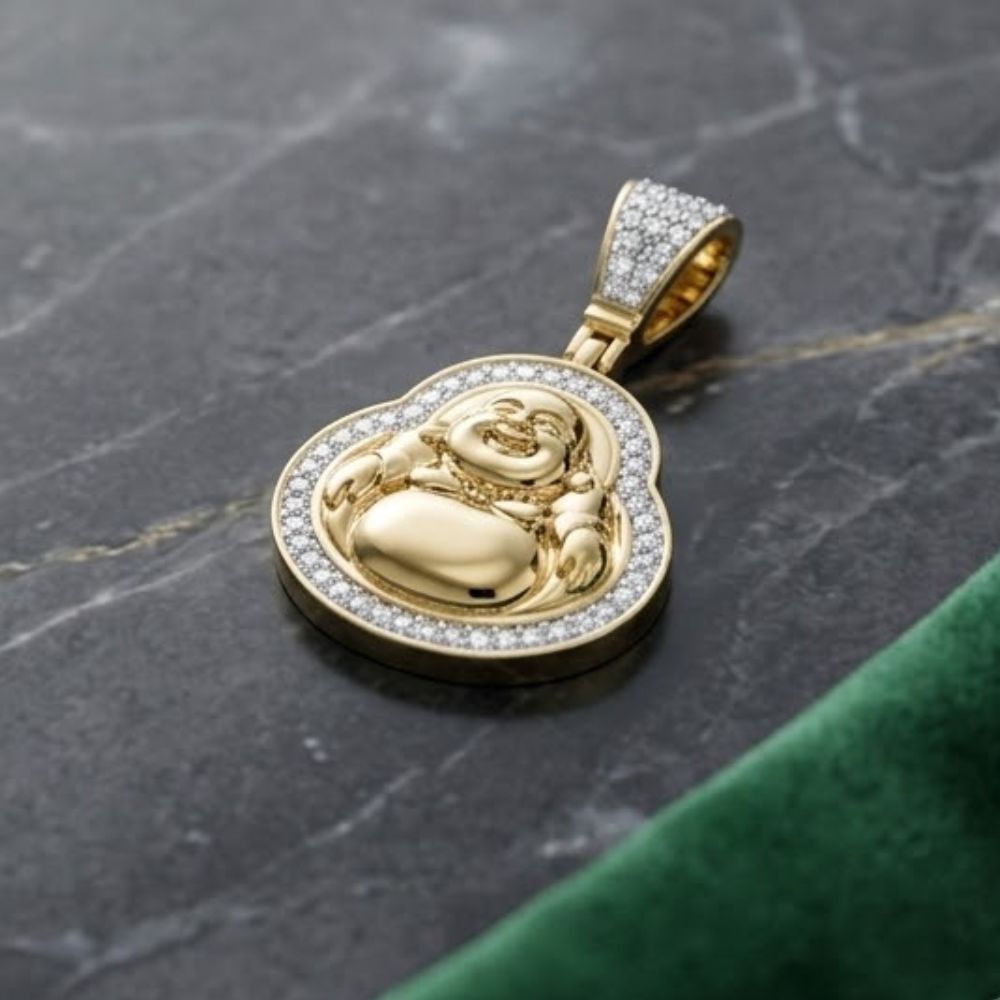 The Diamond Buddha Charm 1/3CT in 10K yellow gold, crafted in the shape of a laughing Buddha and set with sparkling diamonds, is displayed on dark marble next to green velvet fabric.