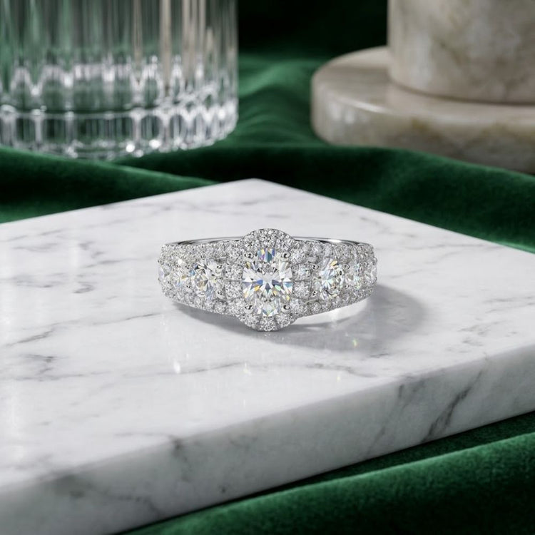 A captivating 14k White Gold Oval Diamond Halo Engagement Ring, featuring a prominent oval center stone surrounded by a halo of smaller diamonds on the band, rests gracefully atop a white marble slab, with a green velvet cloth and glassware providing an elegant backdrop.