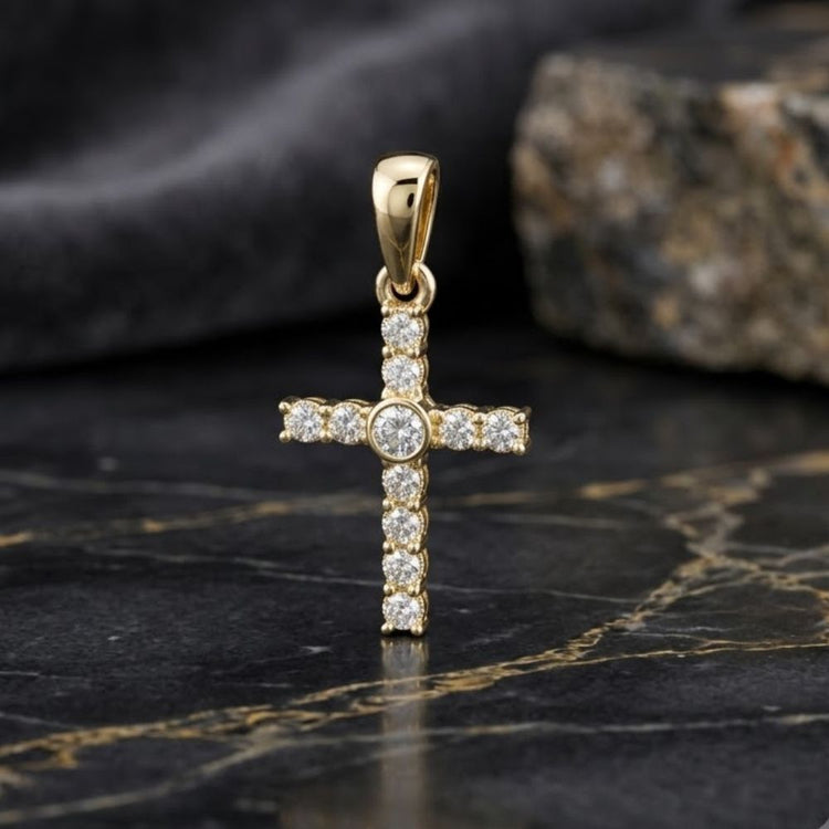 The 10K Yellow Gold 1/20 CTW Diamond Cross Pendant stands upright on a black marble surface, its polished gold gleaming beneath an array of sparkling diamonds, set against a softly blurred stone backdrop.