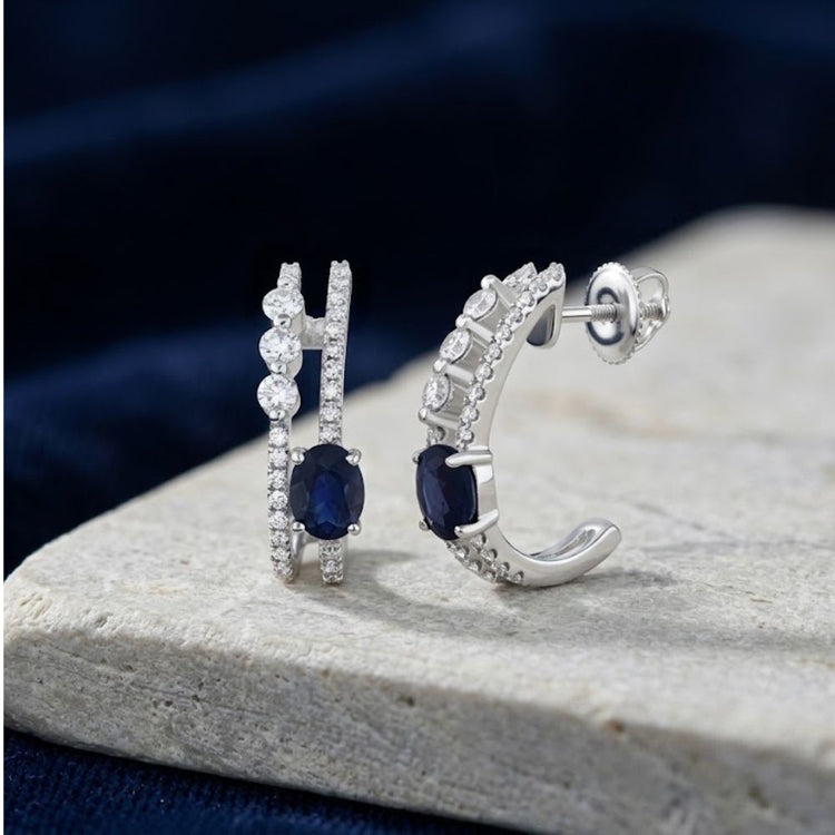14K White Gold Blue Sapphire Hoop Earrings featuring two oval blue sapphires, each flanked by small clear stones on a double-band design.