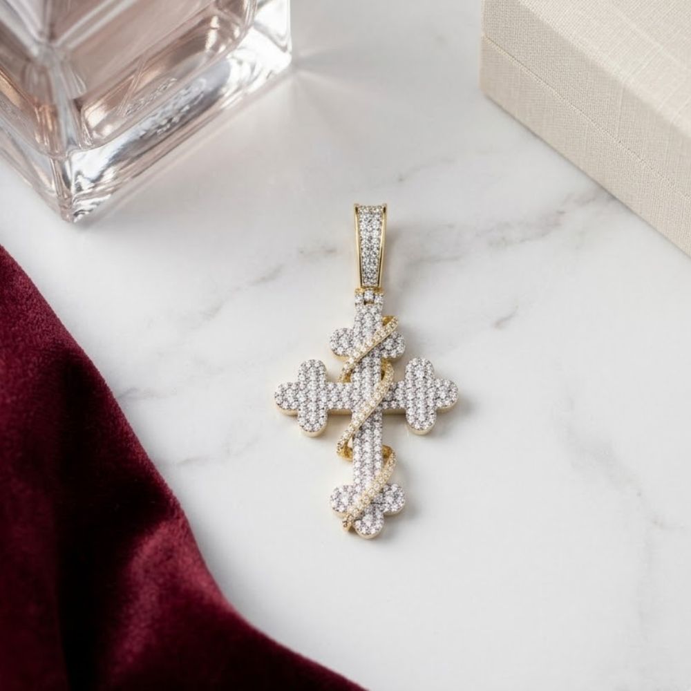 The Diamond Cross Charm 3/4CT 10K Yellow Gold rests on a white surface beside a burgundy cloth, beige box, and glass perfume bottle.