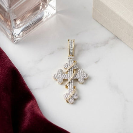 The Diamond Cross Charm 3/4CT 10K Yellow Gold rests on a white surface beside a burgundy cloth, beige box, and glass perfume bottle.