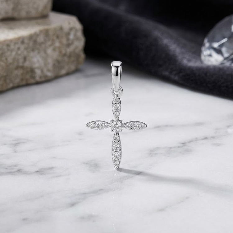 The 10K White Gold Round Diamond Cross Pendant 1/10 CTTW rests on a marble surface, its delicate diamonds sparkling, with a blurred stone block and black fabric in the background.