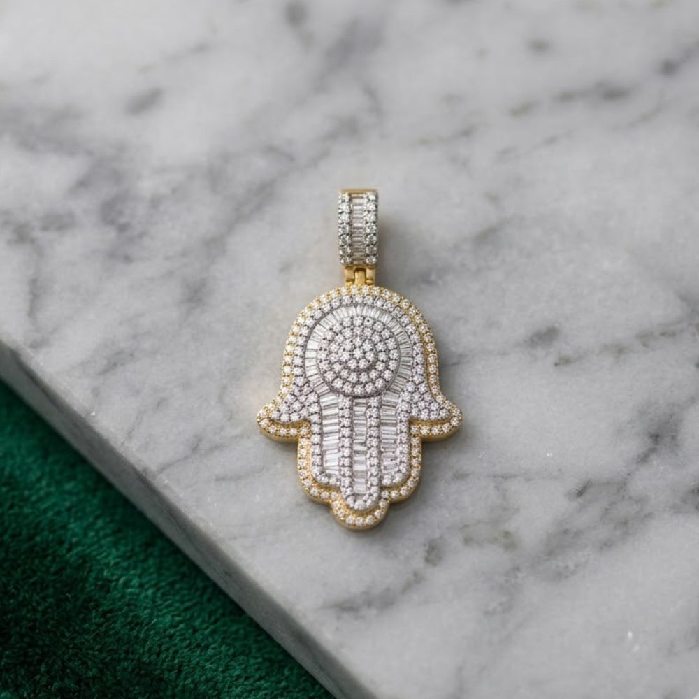 The Diamond Hamsa Pendant 1CT 10K Yellow Gold, adorned with diamonds, is displayed on a white marble surface beside a textured green accent.