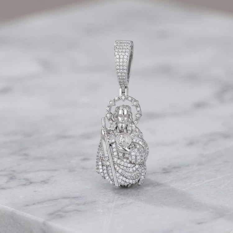 A 925 sterling silver Classic Moissanite St. Jude Charm Pendant, featuring a depiction of the saint holding a child, stands upright on a white marble surface—a striking example of spiritual jewelry.