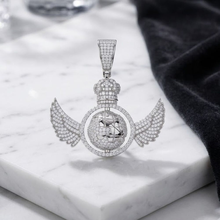 A Sterling Silver Moissanite Rotating Globe Pendant, designed with wing and crown details, rests on a white marble surface next to a black cloth.