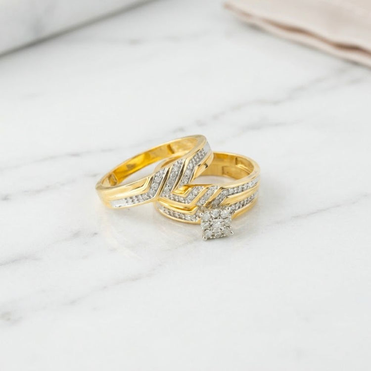 Resting on a white marble surface, the 1/2 CTW 10k Yellow Gold Wedding Trio Set showcases two gold rings adorned with embedded diamonds—one highlighted by a square diamond cluster and the other featuring an elegant zigzag design—making this set a stunning addition to any diamond wedding rings collection.