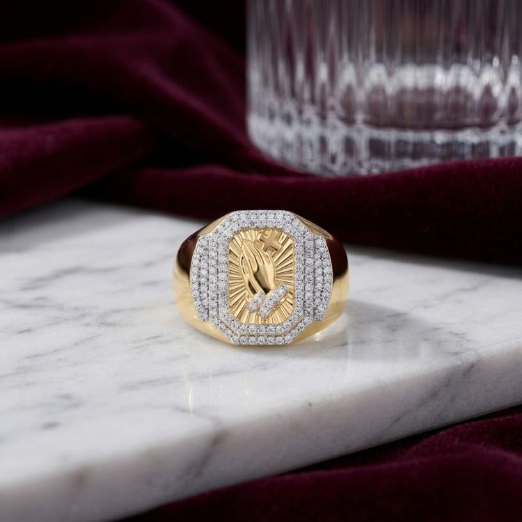 A 1/2CTW Diamond Praying Hands Cross Men’s Ring in 10k gold is displayed on a white marble surface, with a glass and burgundy velvet fabric in the background.