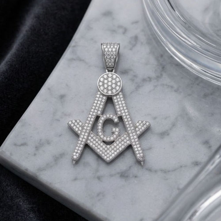 A stunning Masonic pendant, expertly crafted in sterling silver and adorned with diamond-studded details, takes the form of the iconic Freemason square and compass symbol with a prominent G in the center, resting elegantly on a marble surface.