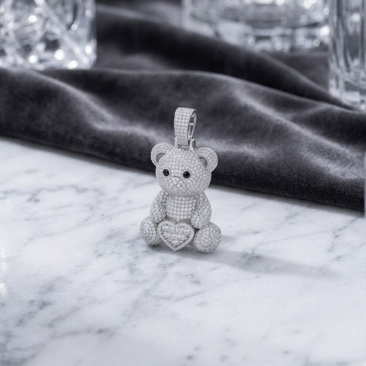 The Sterling Silver Moissanite Teddy Bear Heart Pendant, featuring shimmering black and white moissanite accents and a sparkling heart detail, is elegantly displayed on a marble surface with dark velvet cloth and crystal glasses in the background.