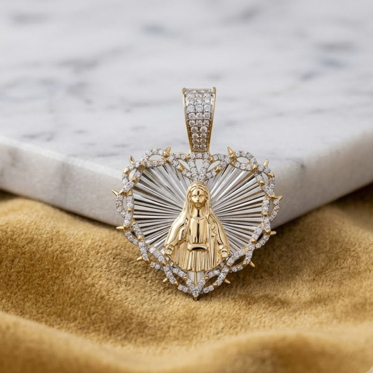 The 3/4 CTW Diamond Mother Mary Heart Charm in 10K gold, featuring sparkling diamonds and a raised Virgin Mary design, is displayed on soft gold fabric next to a white marble surface.