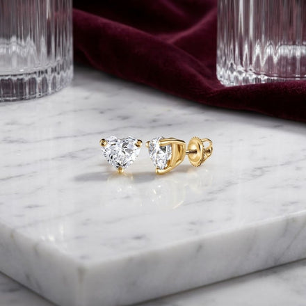 Two 1/5 CTW Natural Diamond Heart Stud Earrings with gold settings rest on a white marble surface, beside clear glass tumblers and a piece of dark red fabric in the background.