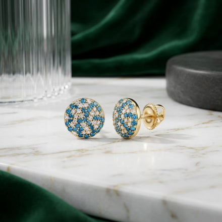 The 5/8CTW Blue Diamond Earrings in 10K Yellow Gold, featuring blue and clear gemstones, are displayed on a white marble surface. A glass object and green fabric in the background lend an extra layer of sophistication to these elegant natural diamond studs.