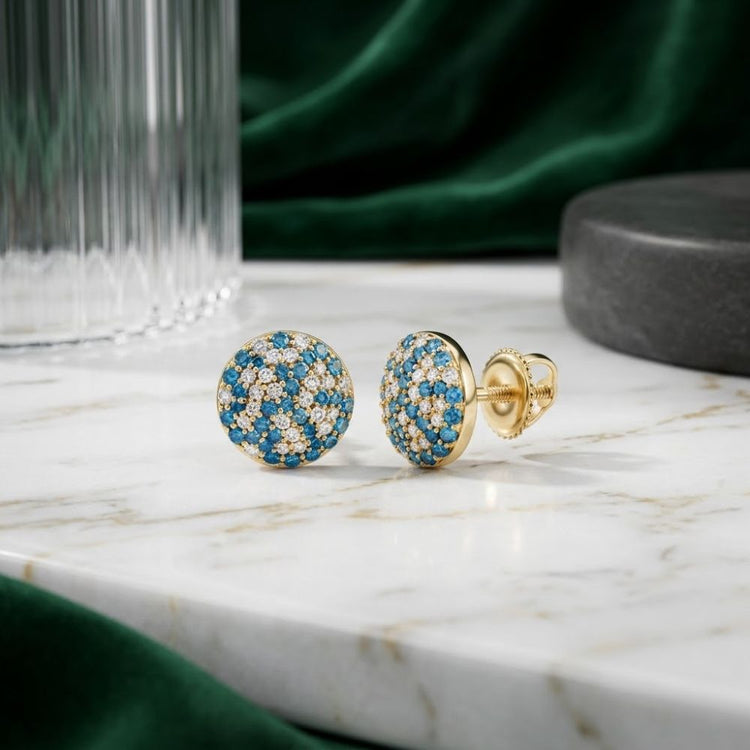 The 5/8CTW Blue Diamond Earrings in 10K Yellow Gold, featuring blue and clear gemstones, are displayed on a white marble surface. A glass object and green fabric in the background lend an extra layer of sophistication to these elegant natural diamond studs.