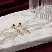 Two 10K yellow gold 1/2 CTW Natural Diamond Paper Clip Earrings, featuring red gemstone studs and dangling oval links each accented with small natural diamonds, rest on a white marble surface with glass decor and a burgundy fabric background—these elegant earrings showcase a contemporary paper clip design.