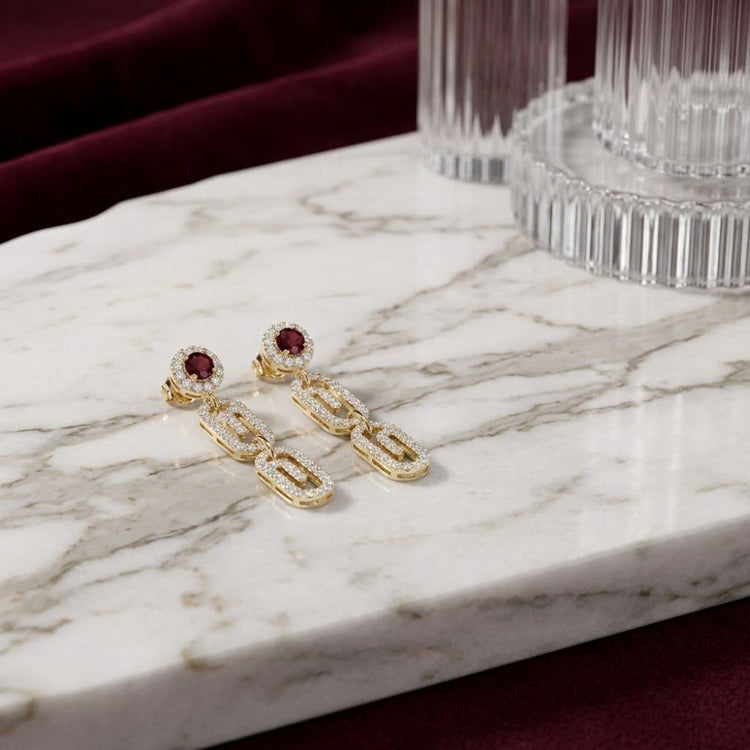 Two 10K yellow gold 1/2 CTW Natural Diamond Paper Clip Earrings, featuring red gemstone studs and dangling oval links each accented with small natural diamonds, rest on a white marble surface with glass decor and a burgundy fabric background—these elegant earrings showcase a contemporary paper clip design.