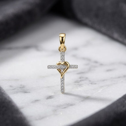 A 10k Yellow Gold Diamond Heart Cross Pendant 1/20 Cttw, featuring a radiant gold heart at its center and sparkling diamonds, rests elegantly on a white marble surface framed by dark gray fabric.