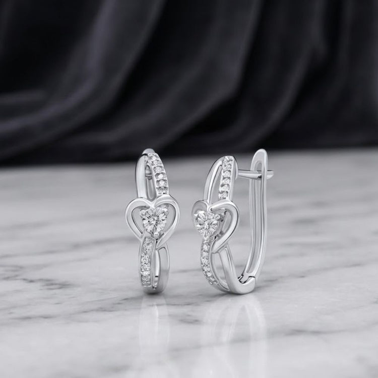 The Sterling Silver Diamond Heart Hoop Earrings, featuring intricate heart-shaped designs accented with small diamonds, are displayed on a marble surface against a softly blurred dark fabric background.