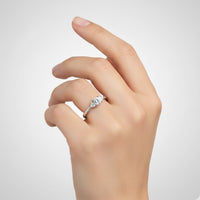 A hand with neatly manicured nails is shown wearing the Round Brilliant Cut Diamond Engagement Ring—featuring a single large round brilliant cut diamond set in silver—on the ring finger, against a plain light gray background.