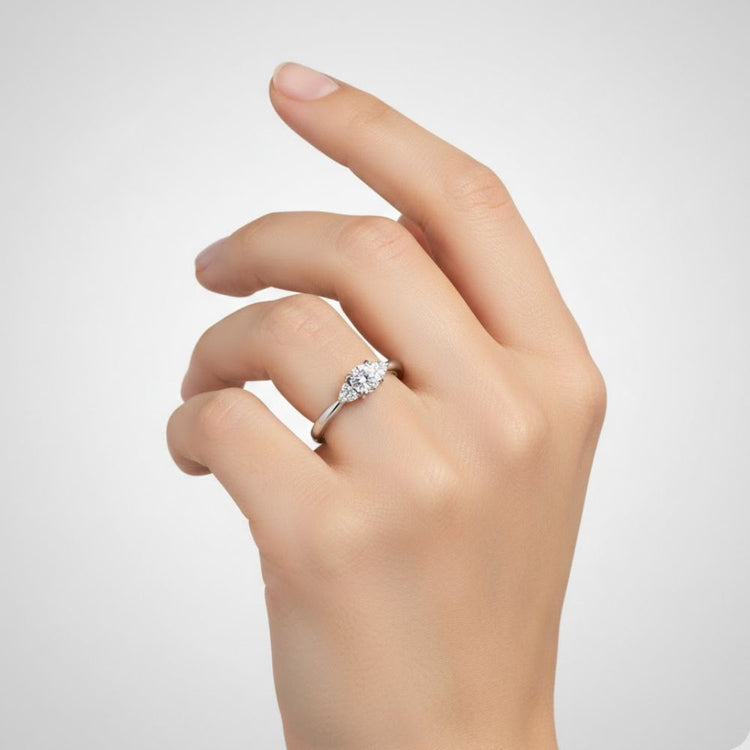 A hand with neatly manicured nails is shown wearing the Round Brilliant Cut Diamond Engagement Ring—featuring a single large round brilliant cut diamond set in silver—on the ring finger, against a plain light gray background.