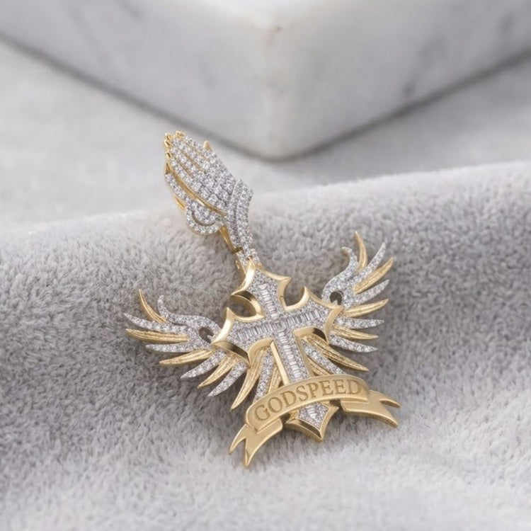 The Diamond Winged Cross Charm 1 7/8 CTTW Gold is a men's 10K yellow gold and diamond pendant with praying hands above a cross, angel wings, and a "GODSPEED" banner. It is shown resting on gray fabric.