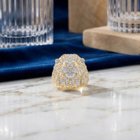 The 1 3/4CTW Lion Face Diamond Men’s Ring in 10k Gold, featuring a meticulously crafted lion face adorned with dazzling diamonds, rests elegantly on a marble surface beside sophisticated glassware and lush blue velvet fabric in the background. This natural diamond men’s ring embodies an aura of luxury and bold power, making it the ultimate statement accessory.