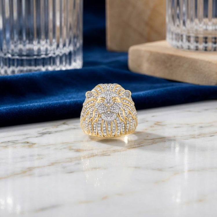 The 1 3/4CTW Lion Face Diamond Men’s Ring in 10k Gold, featuring a meticulously crafted lion face adorned with dazzling diamonds, rests elegantly on a marble surface beside sophisticated glassware and lush blue velvet fabric in the background. This natural diamond men’s ring embodies an aura of luxury and bold power, making it the ultimate statement accessory.